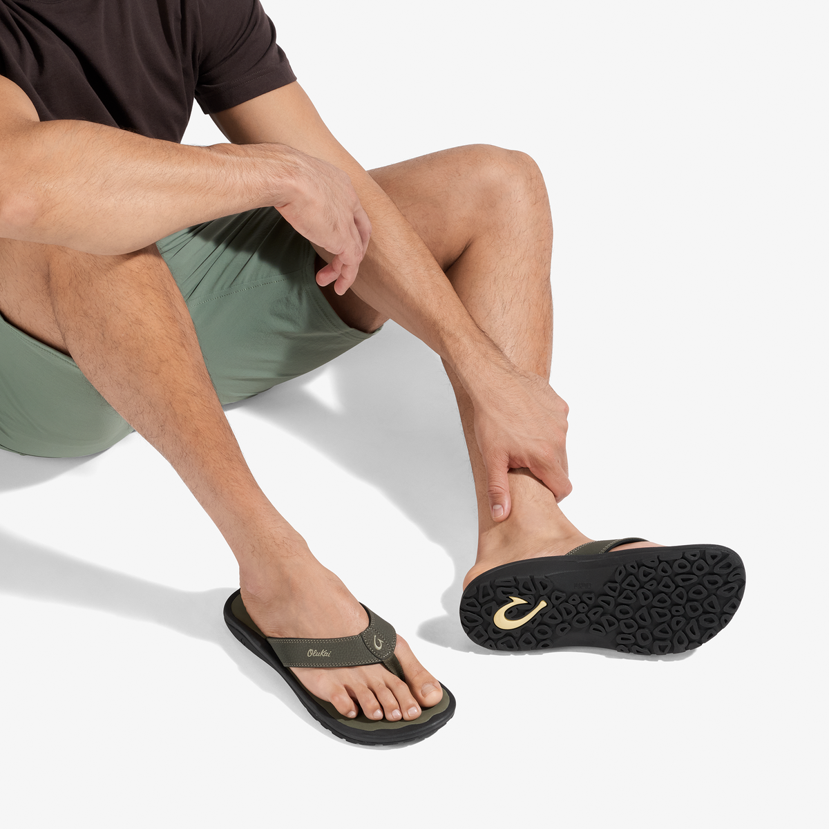 Man wearing sage green shorts and black thong sandal with gold logo detail, shown from waist down.