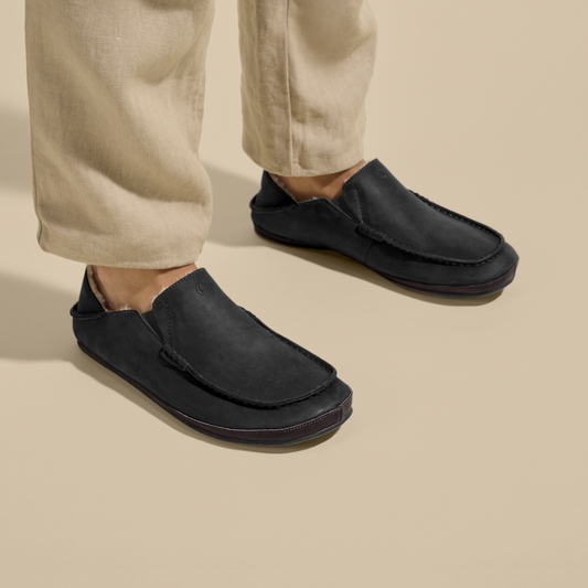 Black suede slip-on slippers with dark brown trim, worn with beige pants.