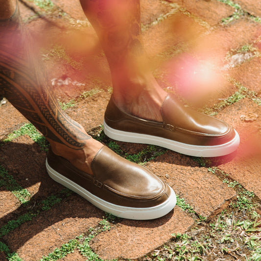 Walnut brown leather slip-on loafers with white sole worn on feet outdoors.