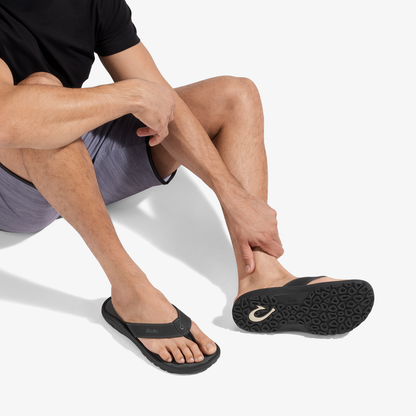 Man wearing black thong sandal with textured sole and dark gray shorts, seated position showing footwear detail.