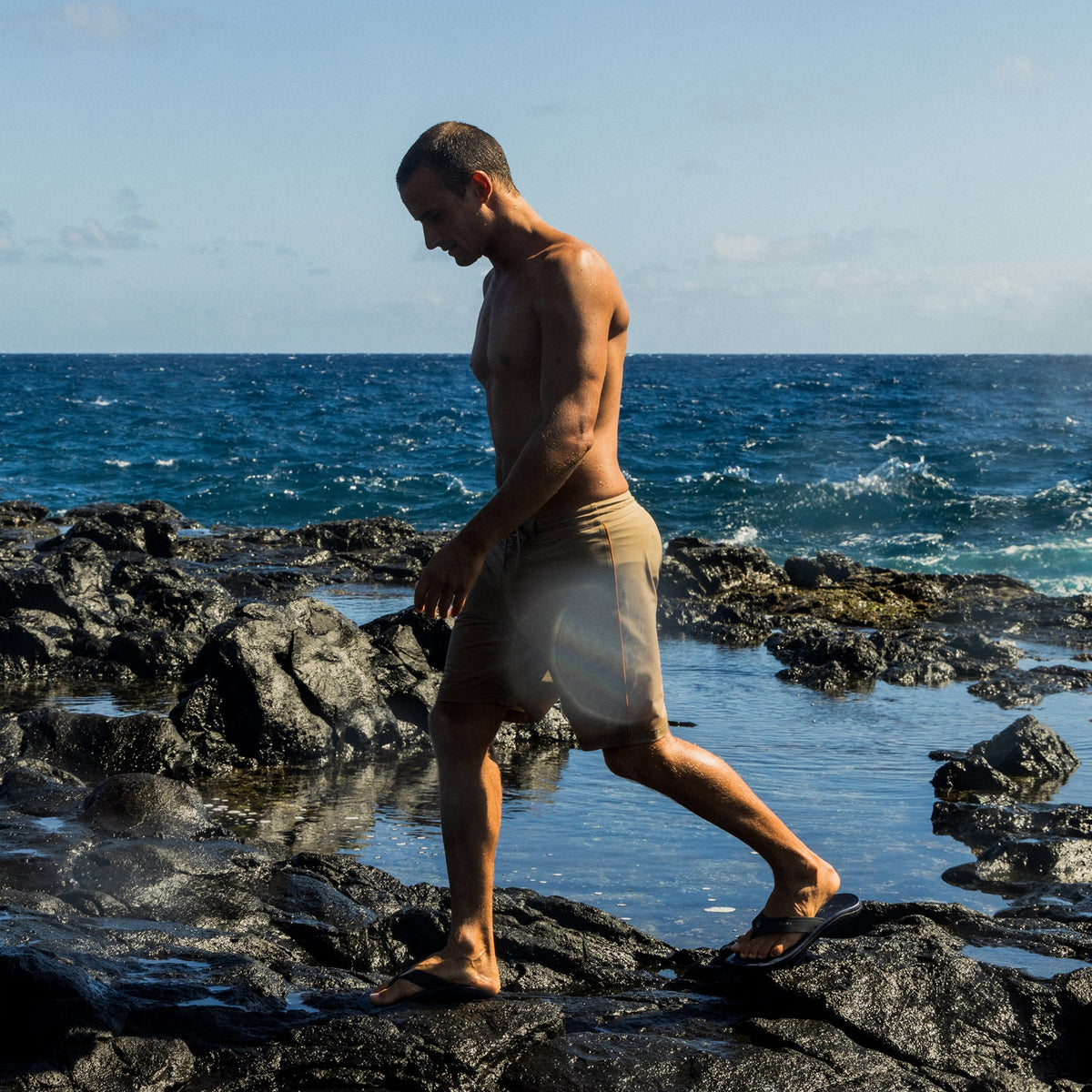 Man wearing trench blue swim shorts standing on rocks by the ocean.