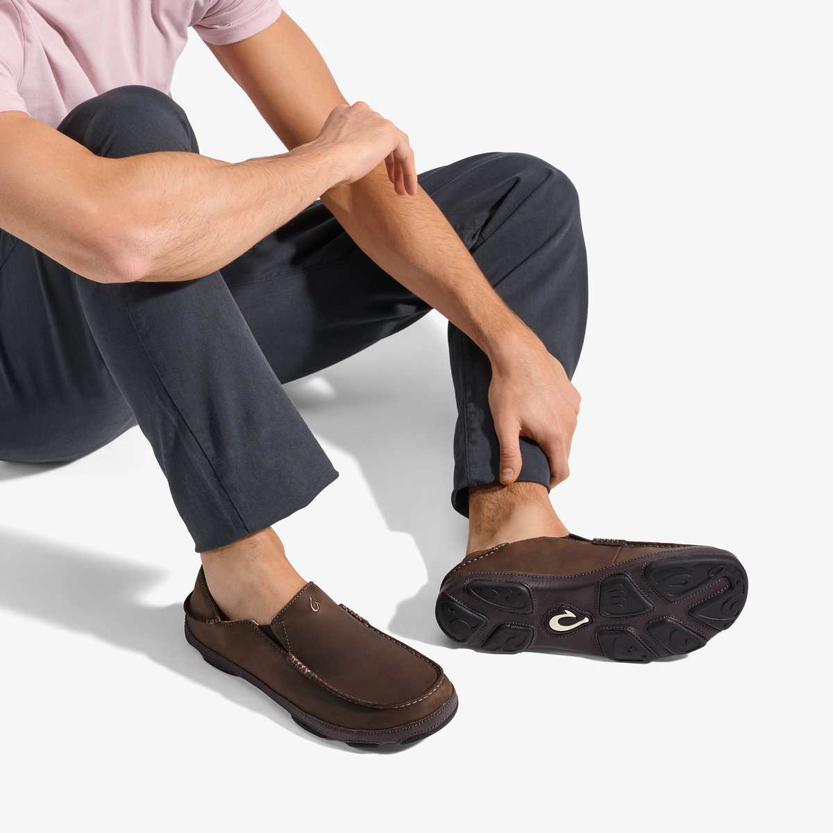 Man wearing dark brown leather slip-on loafer with black sole, navy pants, and pink shirt, shown from seated side view.