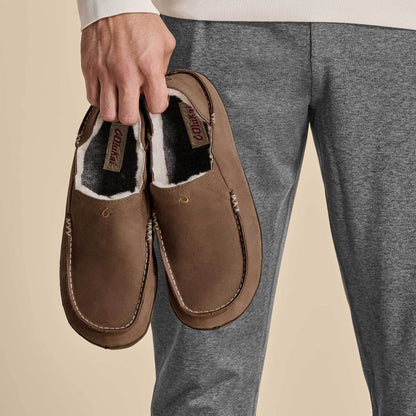 Brown leather slippers with black and white fleece lining, held in hand, showing rounded toe and stitched seams.
