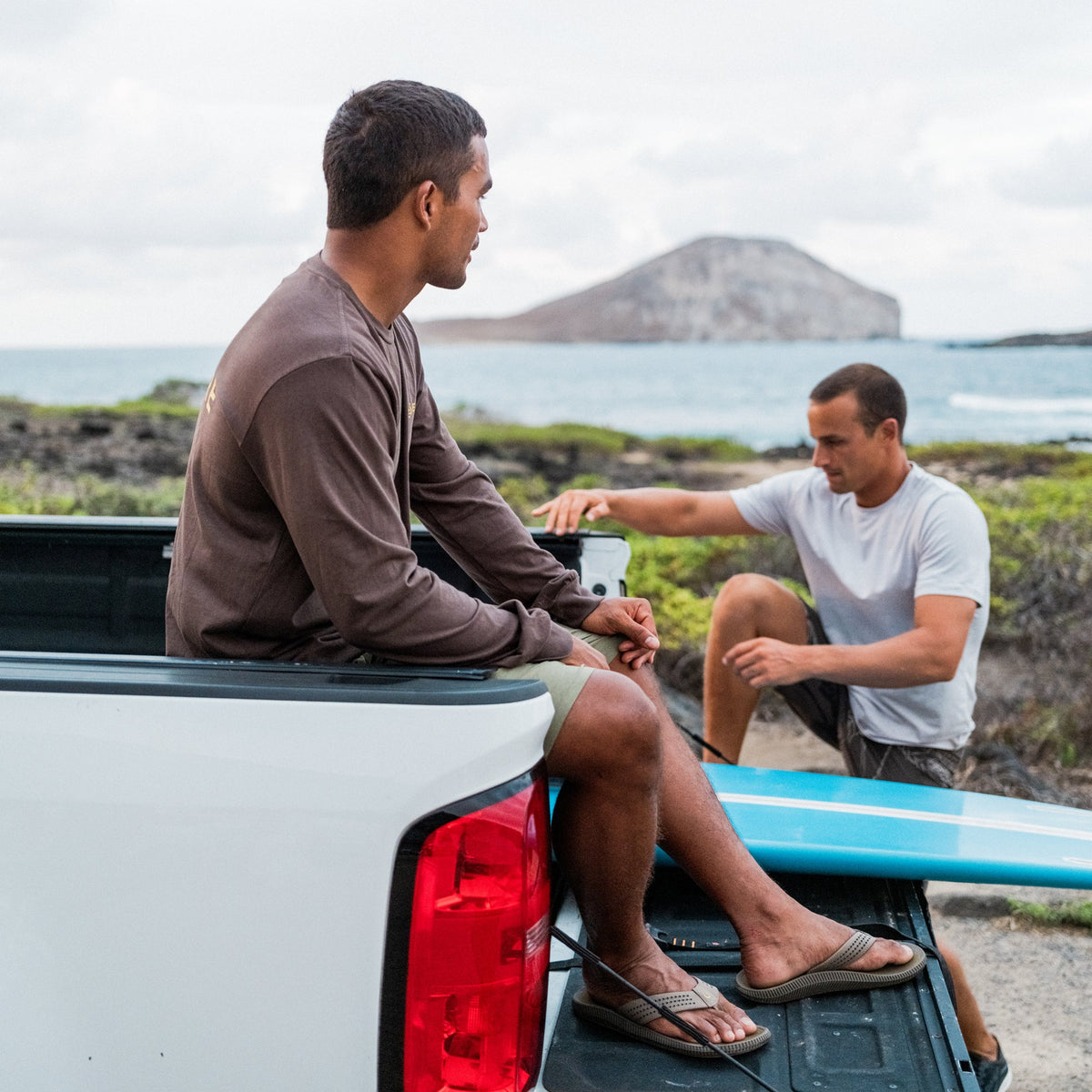 Two men with surfboards on truck bed overlooking coastal landscape with volcanic island.