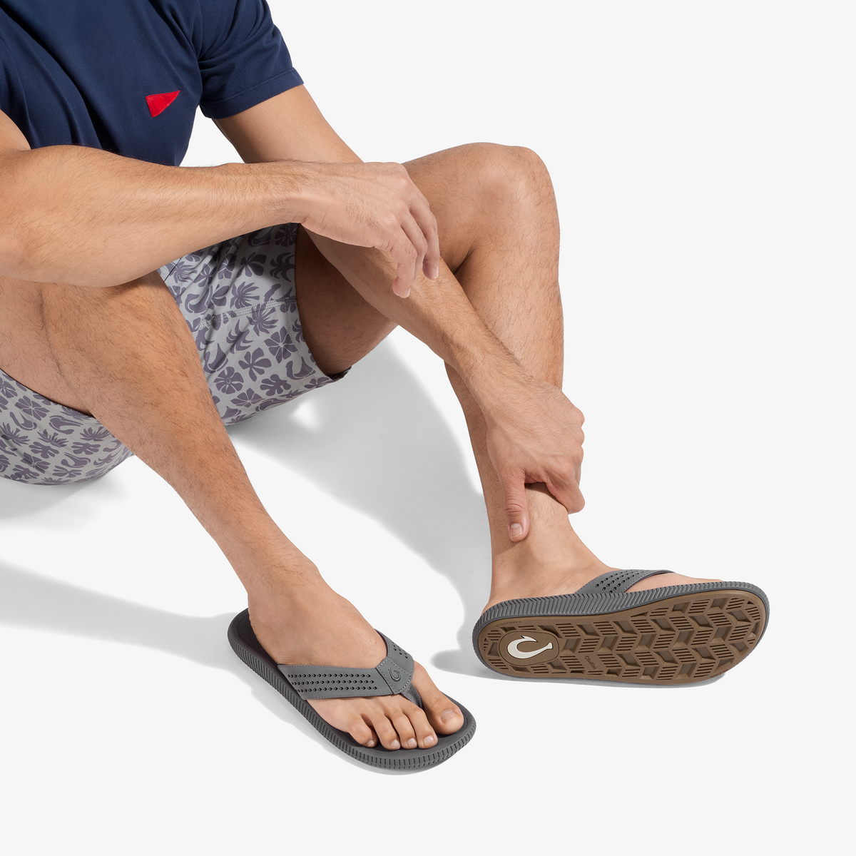 Man wearing navy t-shirt and patterned shorts sitting cross-legged, displaying gray thong sandals with textured soles.