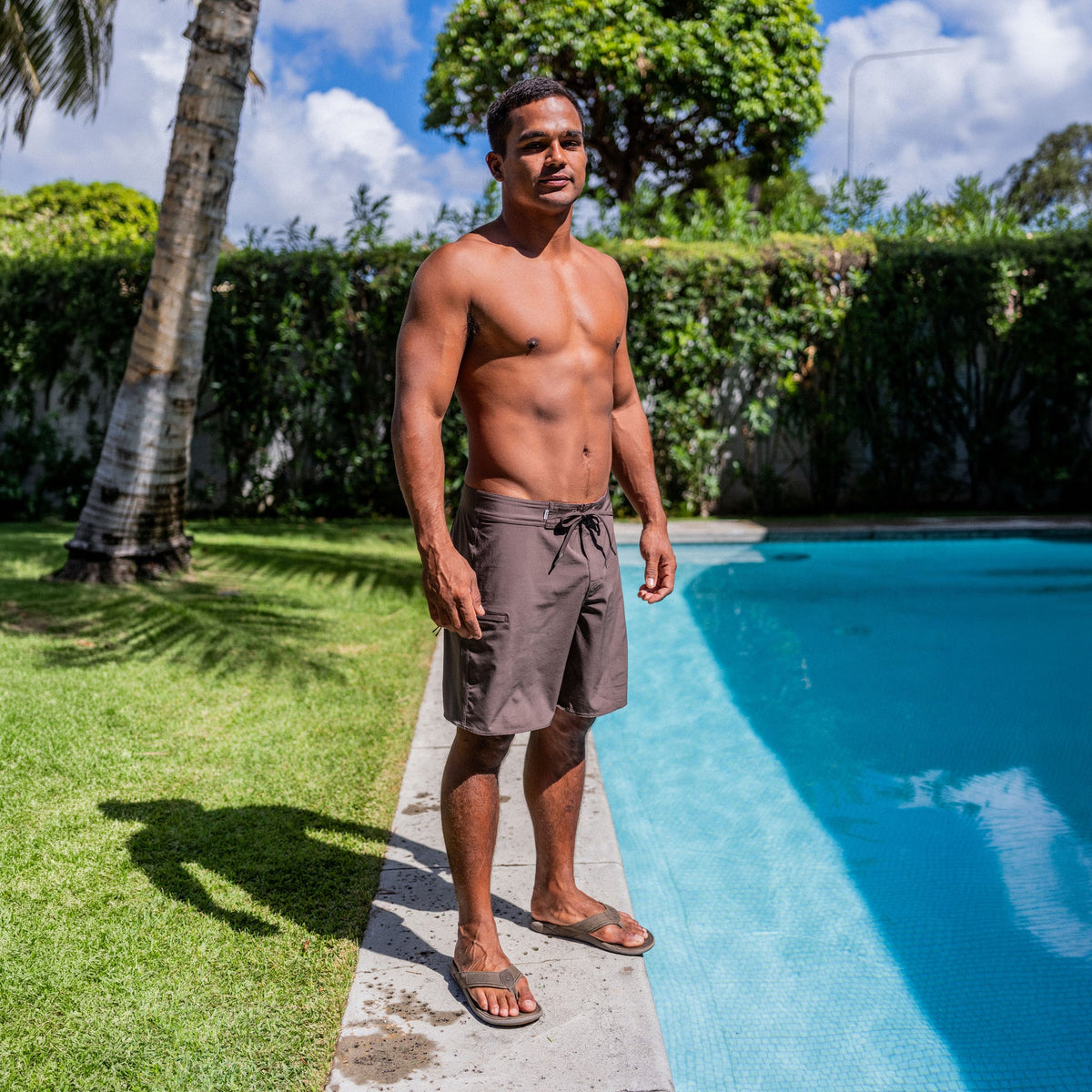 Man wearing gray drawstring shorts and dark slides standing poolside on concrete deck.