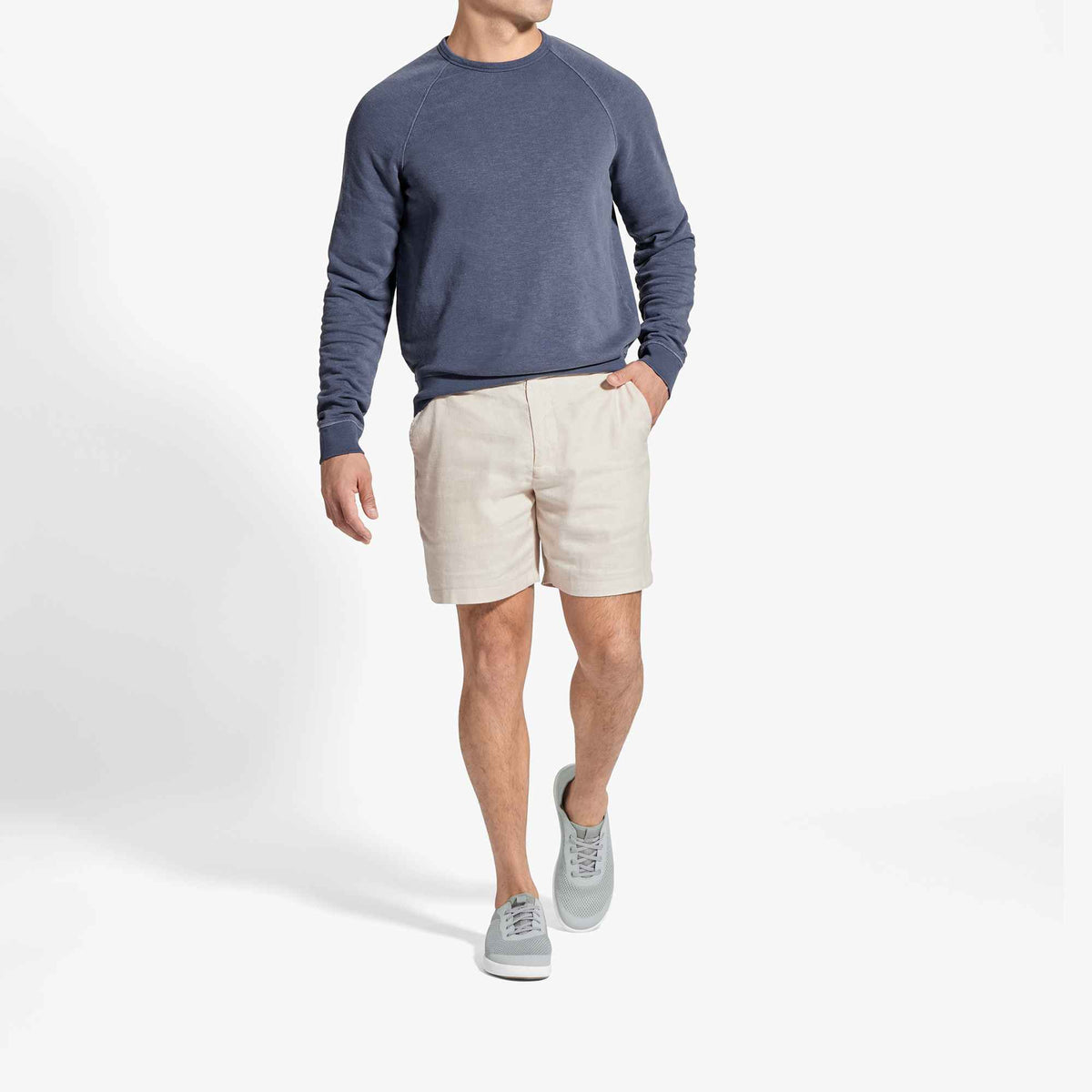 Man wearing blue-grey crewneck sweatshirt with cream shorts and grey sneakers.