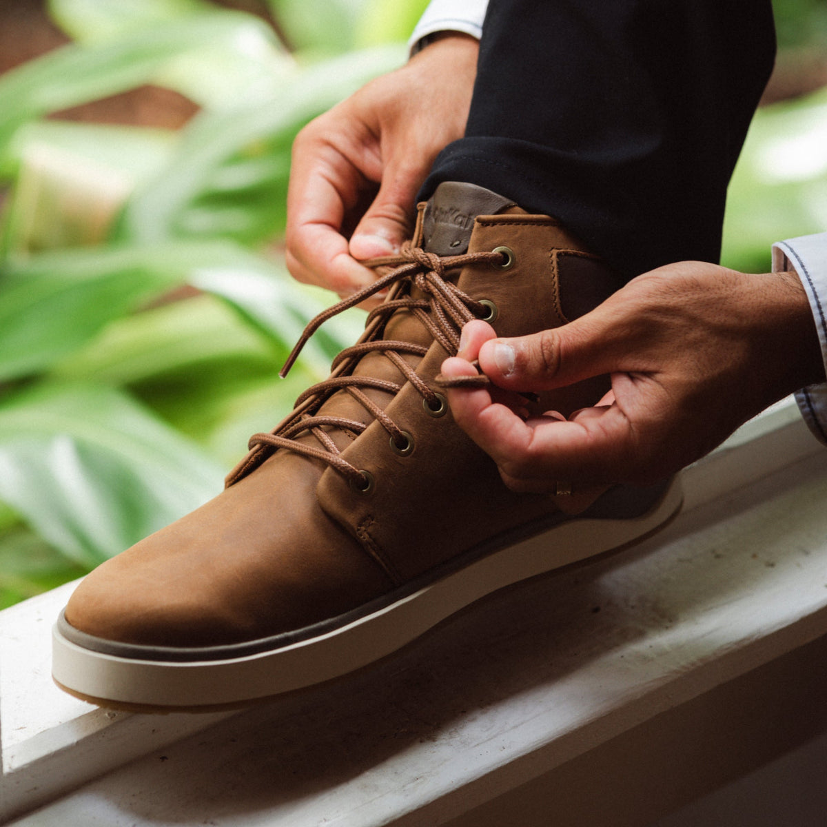 Brown lace-up boot with cream sole being laced by hands, shown on foot outdoors.