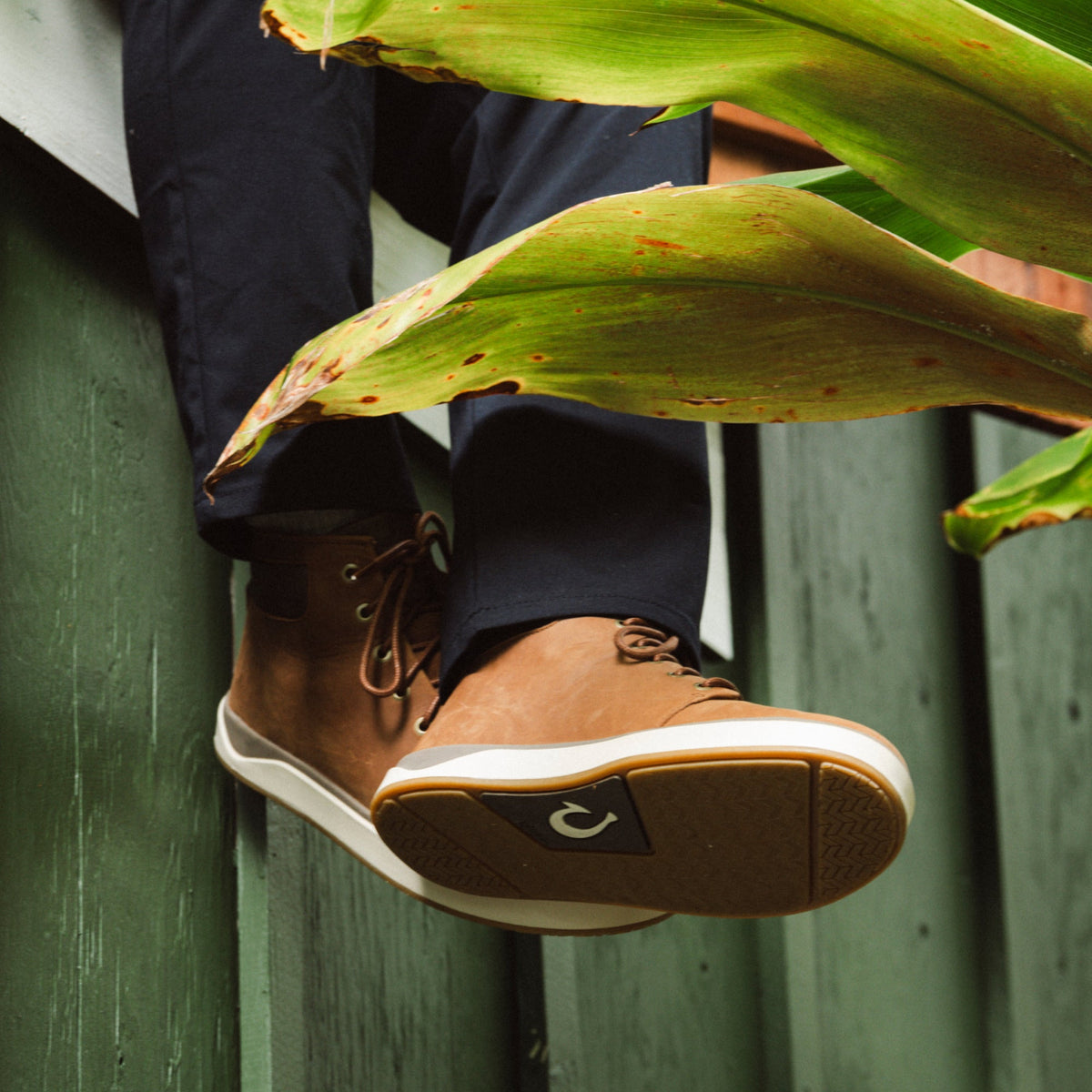 Tan and brown lace-up boot with white sole against green corrugated background and tropical foliage.