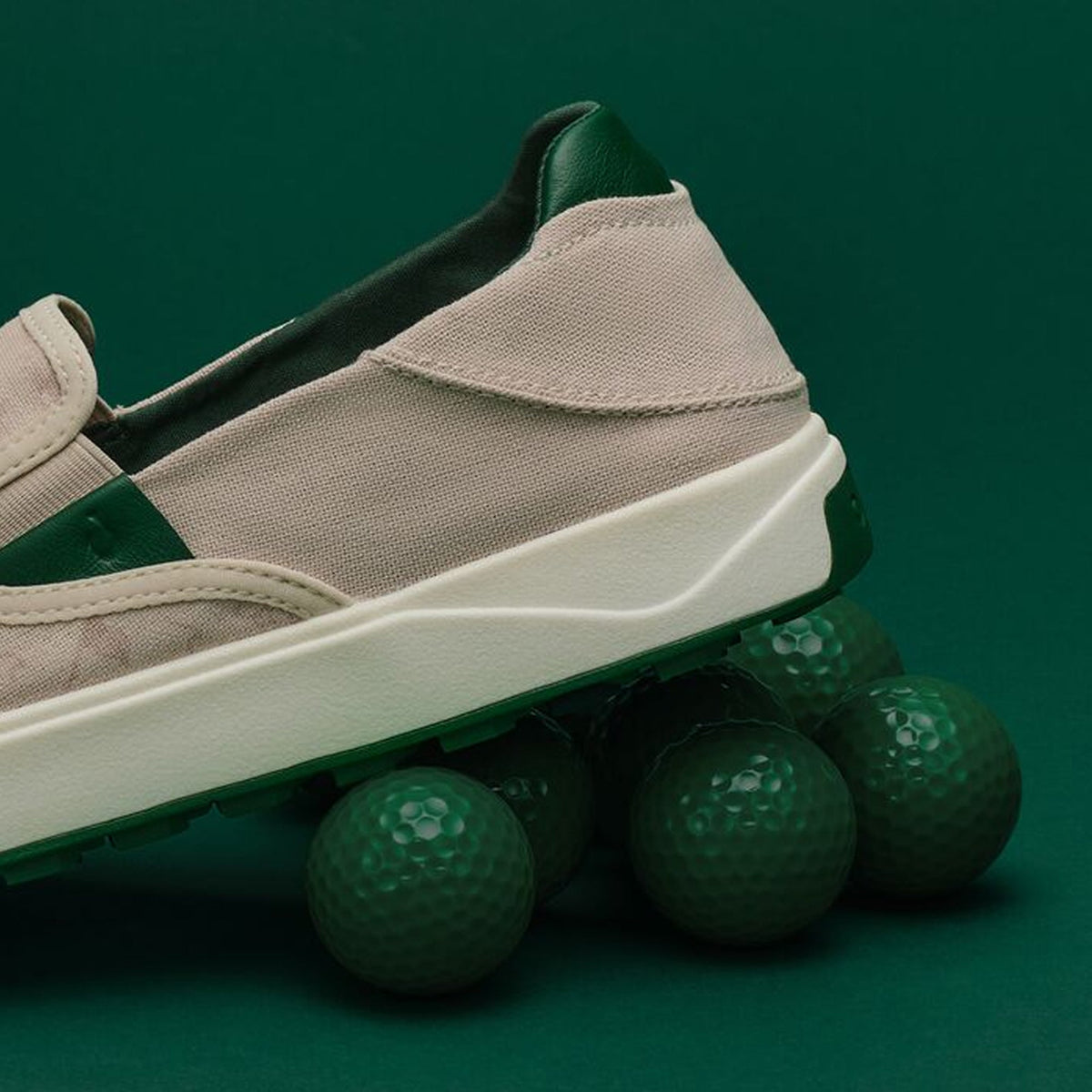 Beige canvas slip-on shoe with cream sole and dark green heel collar, displayed against green golf balls on teal background.