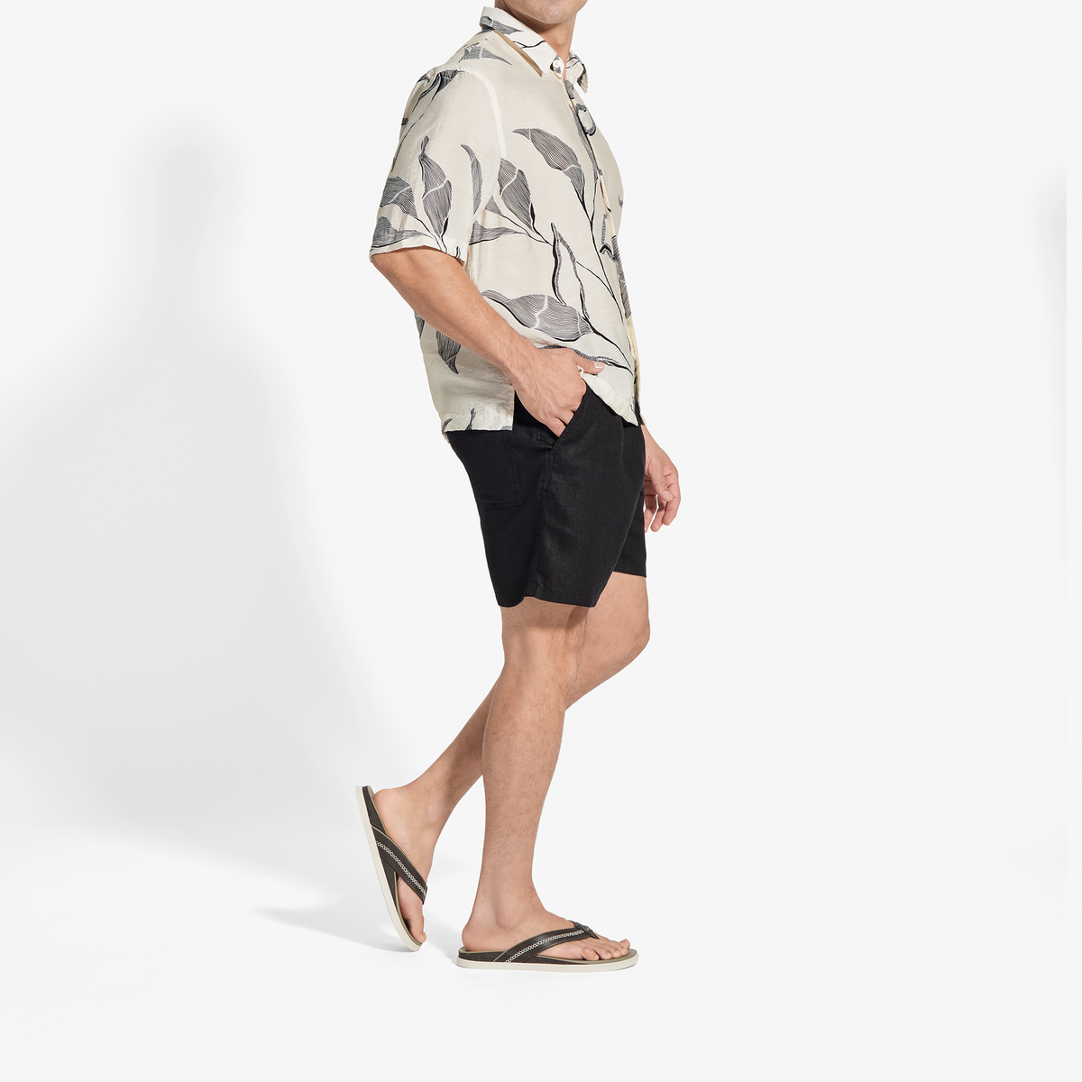 Man wearing cream short-sleeve shirt with gray leaf print and black shorts with sandals.