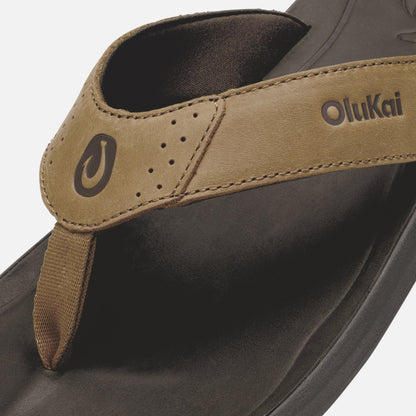 OluKai Pili flip-flop with tan leather strap and dark sole, showing brand logo and perforated detail.
