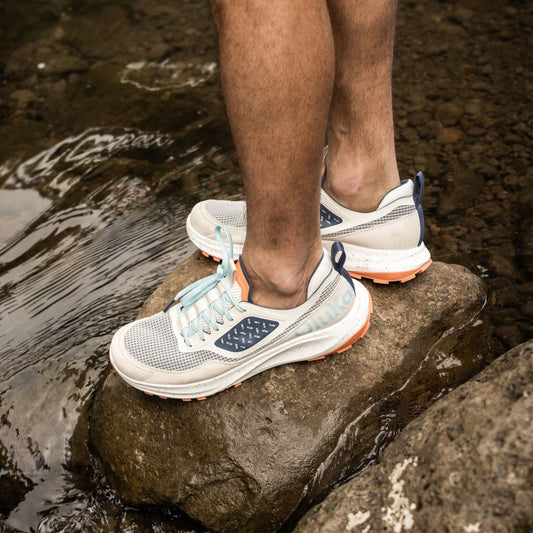 White and navy running shoe with light blue laces worn on person standing on wet rock near water.