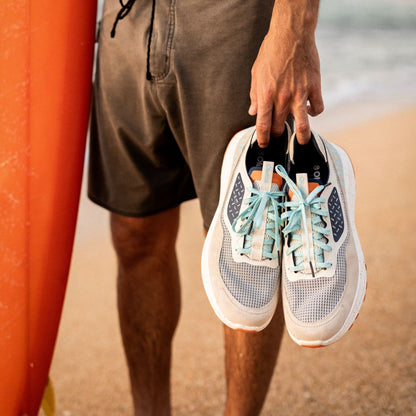 White mesh athletic shoe with mint-green laces held in hand at beach.