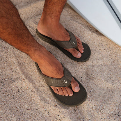 Man wearing brown and black flip-flops with textured straps standing on sandy surface.