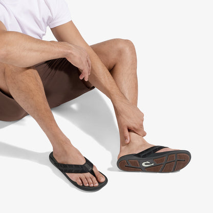 Man wearing white shirt and dark shorts seated, displaying black flip-flop sandal with brown sole detail.