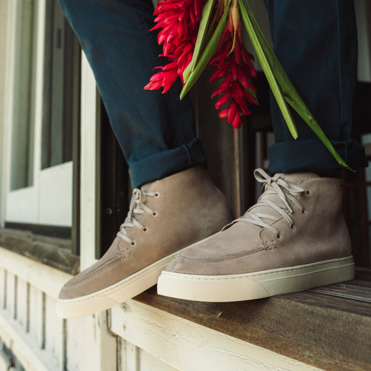 Taupe grey chukka boot with cream sole and white laces, worn with navy pants on a windowsill.