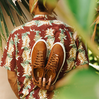 Tan lace-up sneaker with white sole and dark laces, held against a floral burgundy shirt.