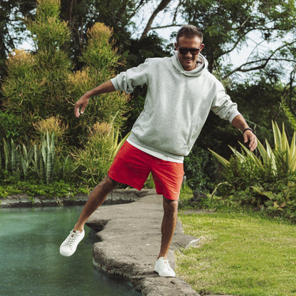 Man wearing white hoodie and red shorts jumping over rocks by a stream with tropical vegetation in background.