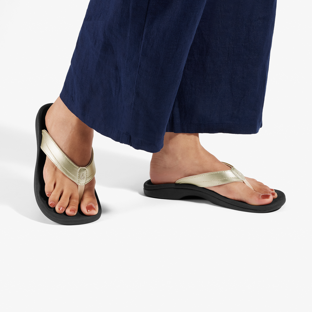 Gold metallic and black thong sandals with cushioned soles worn with navy blue pants.