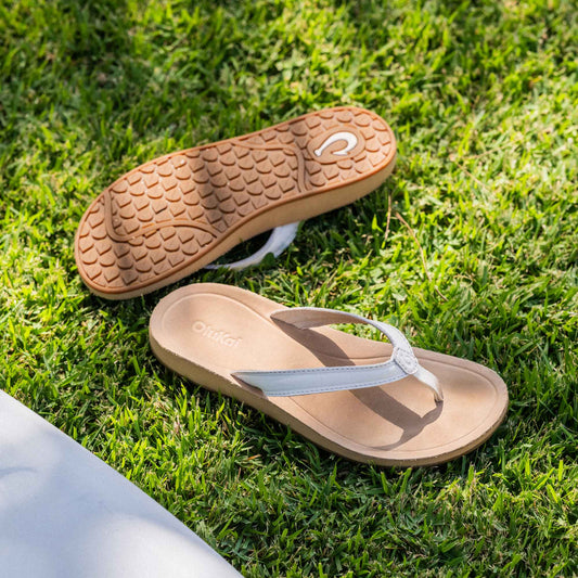 Tan and white flip-flop sandal with textured sole displayed on grass, showing top and bottom views.