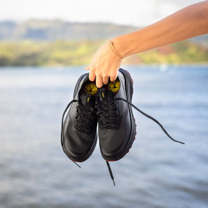 Black lace-up athletic shoes with yellow accents held in hand against water background.