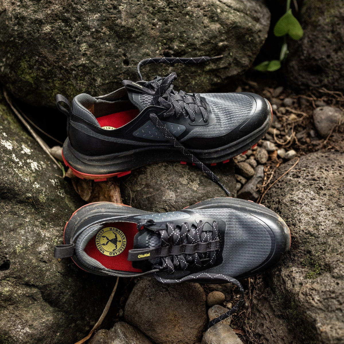 Black and gray trail running shoes with red midsole and accents displayed on lava rock.