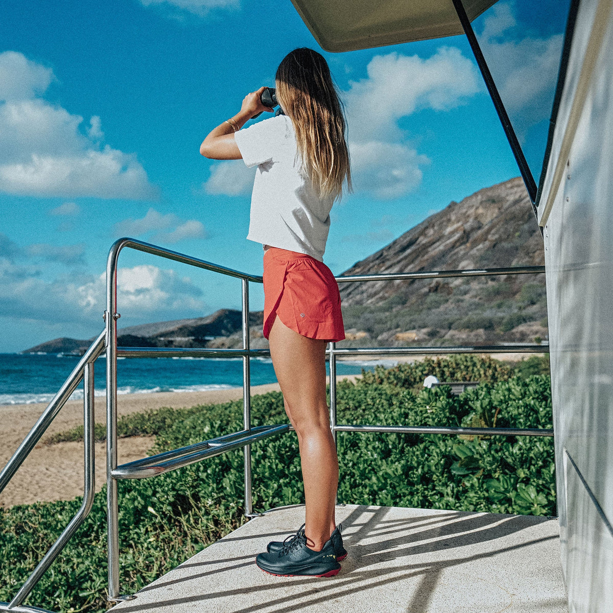 Woman wearing white t-shirt and coral shorts standing on deck overlooking beach and volcanic landscape.