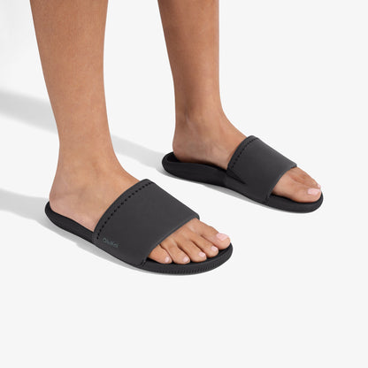 Black slide sandals with textured straps and ribbed soles worn on bare feet.