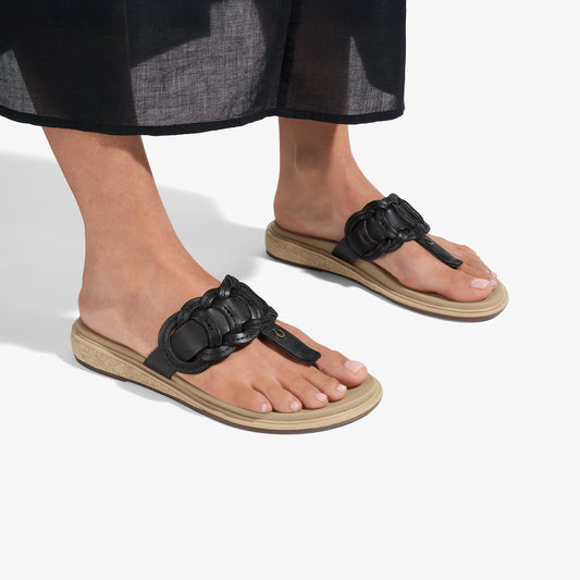 Black ruched thong sandals with beige soles worn on feet with patterned black and gray hem visible above.