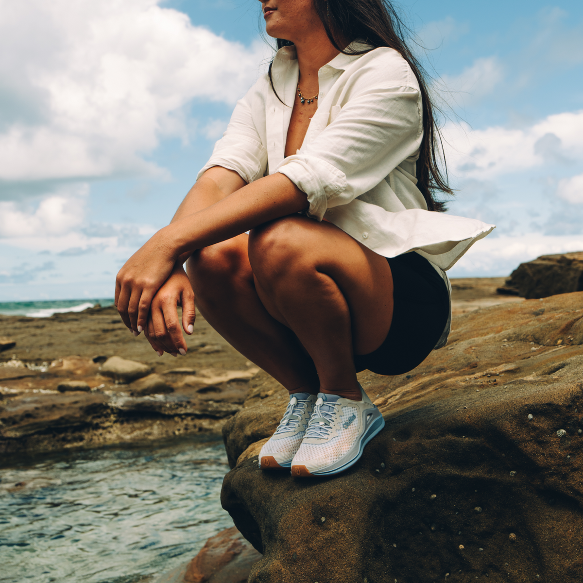 Woman wearing bright white button-up shirt and blue running shoes with teal accents, seated outdoors by ocean.